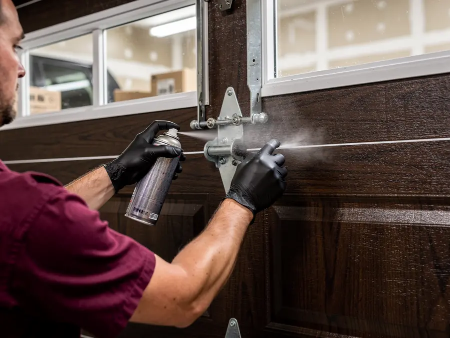 A technician sprays lubricant on a garage door hinge.